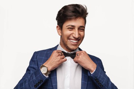 Téléchargez les photos : Heureux jeune homme ethnique en costume élégant avec montre-bracelet de luxe regardant la caméra avec sourire et réglage noeud papillon sur fond gris - en image libre de droit