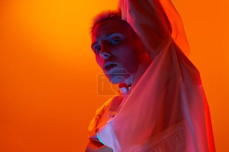 Side view of confident young extravagant female model with short blond hair in translucent coat looking at camera against orange background in neon light