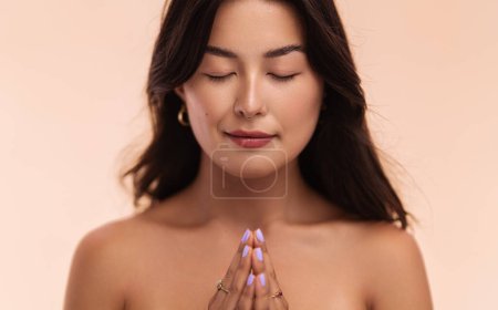 Photo for Peaceful young Asian female with bare shoulders and black hair clasping hands and closing eyes while praying against beige background - Royalty Free Image
