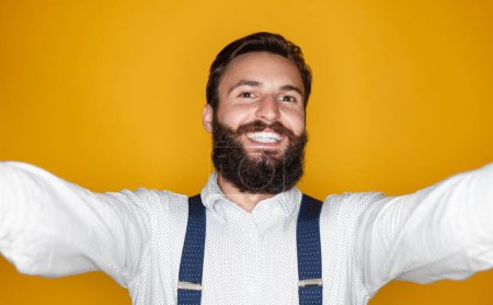 Foto de Hombre barbudo feliz en camisa elegante con tirantes sonriendo y mirando a la cámara mientras toma selfie contra el fondo amarillo - Imagen libre de derechos