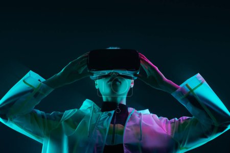 Foto de Mujer joven en impermeable futurista tocando casco VR y explorando el ciberespacio bajo una colorida luz de neón sobre un fondo oscuro - Imagen libre de derechos