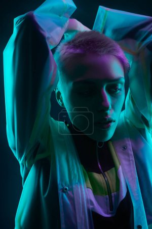 Photo for Androgynous young woman in futuristic raincoat with short hair raising arms under colorful illumination against black background - Royalty Free Image