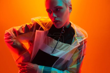 Téléchargez les photos : Jeune modèle féminin androgyne en vêtements futuristes tenant la main à la taille et regardant loin sous un éclairage coloré sur fond orange - en image libre de droit