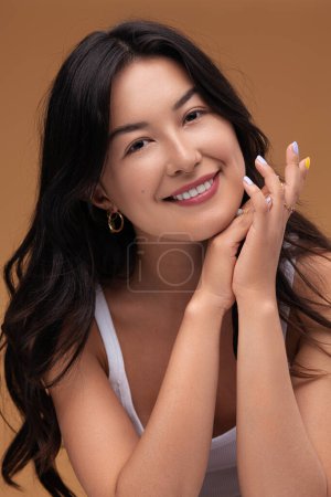 Foto de Mujer asiática feliz con el pelo largo y negro apretando las manos cerca de la barbilla y mirando a la cámara con sonrisa durante la rutina de cuidado de la piel contra el fondo marrón - Imagen libre de derechos