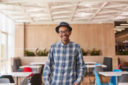 Foto de Feliz joven afroamericano con camisa a cuadros con gafas y sombrero mirando a la cámara y sonriendo alegremente en un amplio espacio de trabajo - Imagen libre de derechos