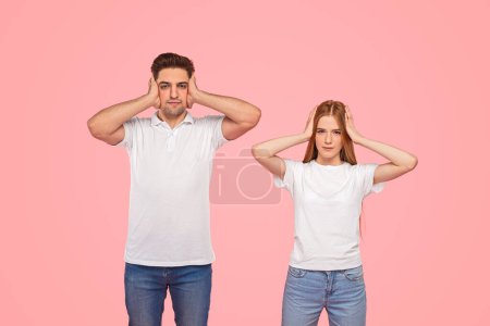 Photo for Unhappy young boyfriend and girlfriend in similar clothes covering ears and looking at camera while ignoring each other after argument against pink background - Royalty Free Image