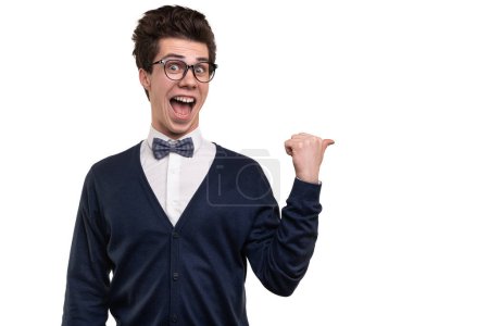 Photo for Funny young man in glasses looking at camera with opened mouth and pointing away with thumb during studies against white background - Royalty Free Image
