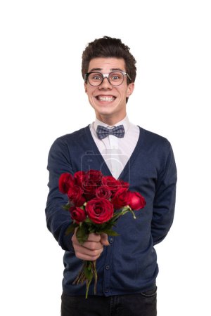 Foto de Feliz joven nerd masculino en ropa casual inteligente y gafas sonriendo y dando rosas rojas a la cámara durante la fecha contra el fondo blanco - Imagen libre de derechos