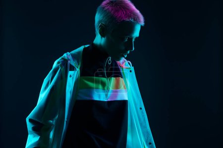 Photo for Young androgynous woman in stylish futuristic clothes with short hair looking away while standing under colorful neon light against black background - Royalty Free Image