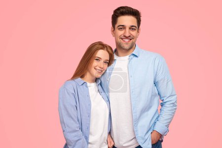 Téléchargez les photos : Jeune homme et femme positive dans des vêtements similaires souriant et regardant la caméra tout en se tenant debout sur fond rose ensemble - en image libre de droit