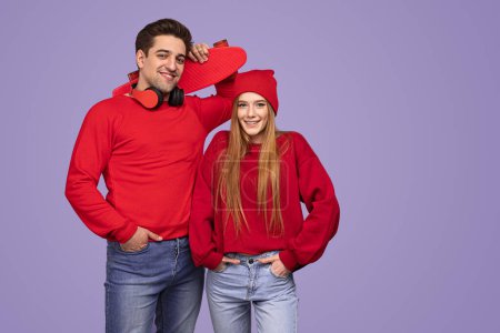 Téléchargez les photos : Positif jeune couple dans des vêtements similaires élégants avec penny conseil tenant la main dans les poches et en regardant la caméra sur fond violet - en image libre de droit
