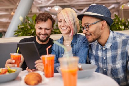Foto de Feliz diversos hombres y mujeres con jugo sonriendo y viendo vídeo en la tableta mientras se sienta en la mesa durante la reunión de amigos en el restaurante moderno - Imagen libre de derechos