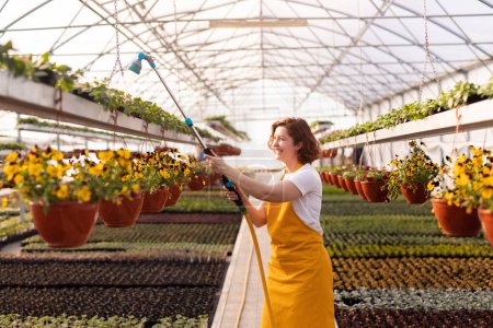 Foto de Mujer alegre en camiseta blanca y delantal amarillo sonriendo y regando plantas en maceta durante el trabajo en invernadero en granjero - Imagen libre de derechos