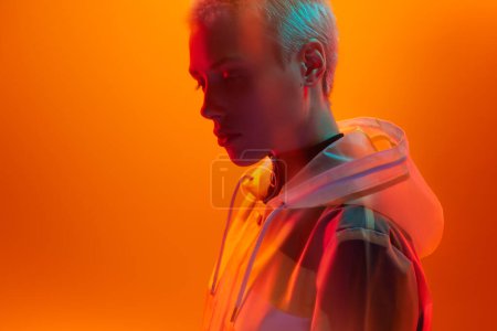 Téléchargez les photos : Jeune modèle féminin androgyne aux yeux futuristes fermant l'imperméable tout en se tenant debout sous un éclairage néon vibrant sur fond orange - en image libre de droit