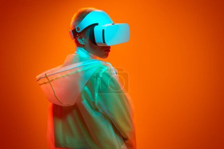 Photo for Young woman in futuristic raincoat and VR headset looking away over shoulder while having virtual reality experience under neon illumination against orange background - Royalty Free Image