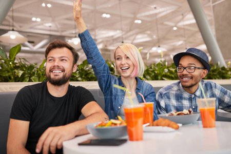 Photo for Happy young female in casual clothes raising arm and greeting friend while sitting between diverse male boyfriends on weekend day in restaurant - Royalty Free Image