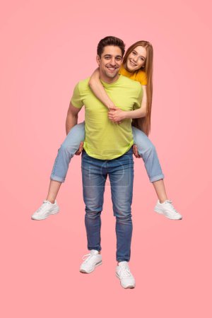 Photo for Full body cheerful young boyfriend in colorful clothes carrying ginger girlfriend on back against pink background - Royalty Free Image