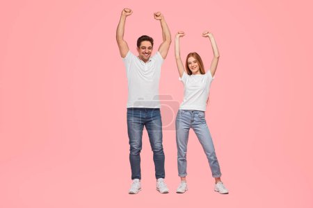 Photo for Full body young boyfriend and girlfriend in similar clothes raising clenched arms and smiling while celebrating victory against pink background - Royalty Free Image