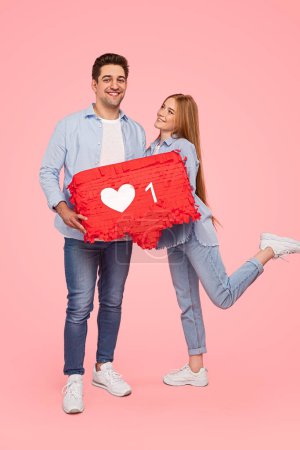 Photo for Full body young man and woman in similar clothes showing speech bubble with like symbol and smiling against pink background - Royalty Free Image