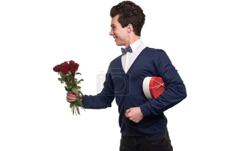 Photo for Happy young man with heart shaped gift box looking away with smile and giving red roses during date against white background - Royalty Free Image