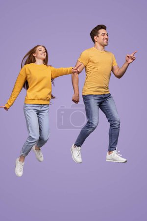 Foto de Cuerpo completo joven hombre y mujer en ropa similar sonriendo y apuntando hacia fuera mientras saltan sobre fondo violeta juntos - Imagen libre de derechos
