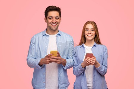 Photo for Cheerful man and woman in similar white tees and blue shirts browsing mobile phones while looking at camera on pink background - Royalty Free Image