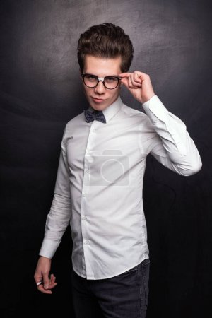 Foto de Hombre joven seguro de sí mismo en ropa casual inteligente haciendo pucheros labios y ajustando gafas contra fondo negro - Imagen libre de derechos