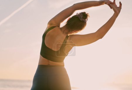 Foto de Back view of anonymous sportswoman raising arms and bending aside against sunset sky during fitness workout near sea - Imagen libre de derechos