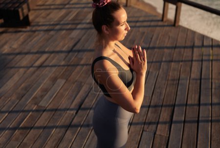 Foto de From above woman in sportswear clasping hands and meditating in Tadasana pose during yoga session on lumber terrace on beach - Imagen libre de derechos