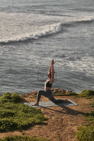 Téléchargez les photos : High angle of slim female raising arms and doing Crescent Lunge on the Knee during yoga session on cliff near waving sea - en image libre de droit
