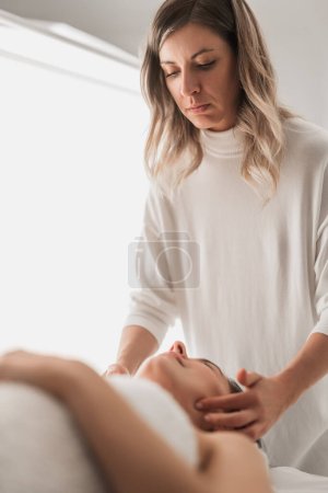 Foto de Professional young female therapist with long blond hair doing face massage to client lying on table during spa session in light salon - Imagen libre de derechos