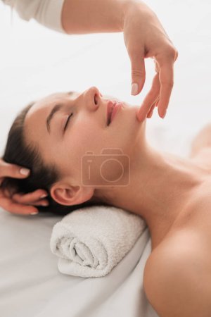 Téléchargez les photos : High angle of peaceful happy young female client lying on table and smiling with closed eyes, while crop cosmetician doing face massage during beauty procedure - en image libre de droit