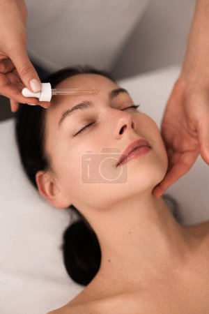 Téléchargez les photos : From above of crop unrecognizable beautician applying essential oil with pipette on face of relaxed young female client with dark hair, lying on table with closed eyes in beauty salon - en image libre de droit