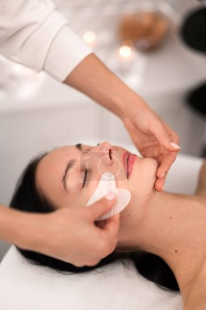 Téléchargez les photos : From above of crop anonymous cosmetician doing face massage with gua sha tool to peaceful young female client with dark hair, lying on couch with closed eyes in salon - en image libre de droit