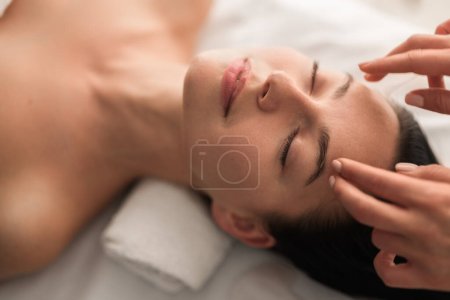Foto de High angle of crop unrecognizable cosmetologist massaging forehead of content young female client with dark hair and perfect skin, lying on table with closed eyes during beauty procedure - Imagen libre de derechos