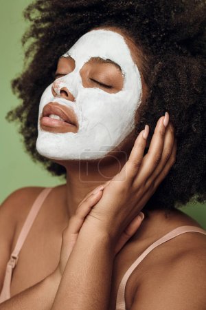 Téléchargez les photos : Calm African American female with white clay mask touching hand and closing eyes during beauty routine against green background - en image libre de droit