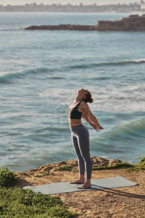 Téléchargez les photos : Side view full body of young barefooted woman in leggings and top stretching arms and back, while standing on mat during yoga session on rocky shore near wavy ocean - en image libre de droit