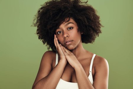 Foto de Young African American female millennial with curly dark hair in white bra touching cheek gently and looking at camera against green background - Imagen libre de derechos