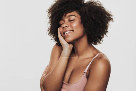 Téléchargez les photos : Cheerful young black female with Afro hairstyle smiling with closed eyes and touching soft skin of clean cheek against gray background - en image libre de droit