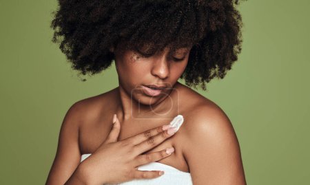 Téléchargez les photos : Young African American female with curly hair applying moisturizing cream on shoulder during spa session against green background - en image libre de droit