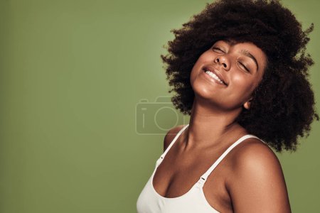 Photo for Side view of confident happy young black female model with dark Afro hair in white bra smiling with closed eyes against green background - Royalty Free Image