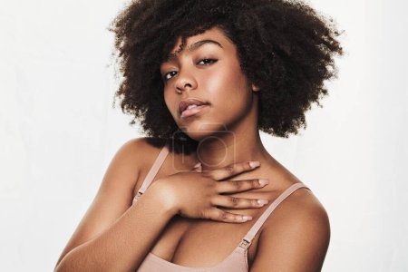 Foto de Self assured young black female model with curly Afro hair in bra touching neck and looking at camera against white background - Imagen libre de derechos