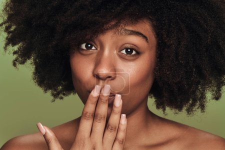 Foto de Confused black female with Afro hairstyle covering mouth and looking at camera after making mistake against green background - Imagen libre de derechos