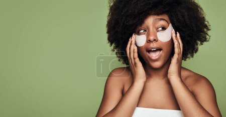 Foto de Positive confident young black female millennial with Afro hair, applying hydrating collagen patches on face and looking away with opened mouth against green background - Imagen libre de derechos