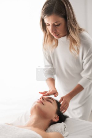 Téléchargez les photos : Concentrated young female cosmetician with long hair applying skin care product on face of client during face massage session in light salon - en image libre de droit