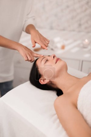 Téléchargez les photos : High angle of relaxed young female client with closed eyes lying on massage table during acupressure facial treatment in modern salon - en image libre de droit