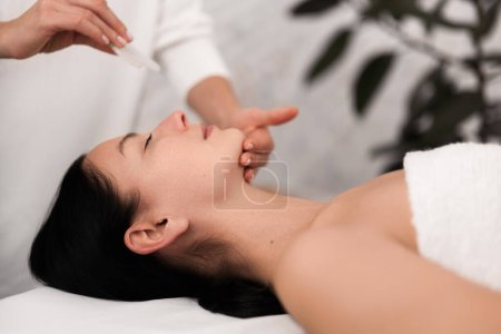 Photo for Side view of young relaxed female client with long dark hair and bare shoulders, lying on table while crop unrecognizable cosmetician doing face massage with gua sha - Royalty Free Image
