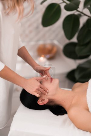 Téléchargez les photos : From above of crop anonymous female beautician with blond hair doing face massage to happy calm woman, lying on table during skincare treatment in spa salon - en image libre de droit
