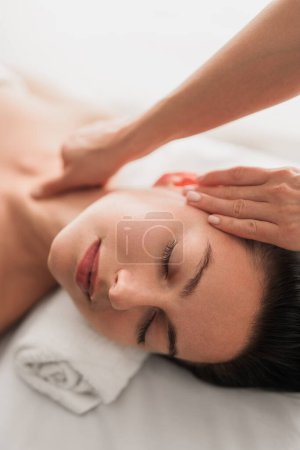 Téléchargez les photos : High angle of young female lying on bed at massage therapy salon. Closeup of face of woman with closed eyes being touched by professional masseur - en image libre de droit