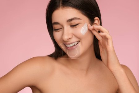 Téléchargez les photos : Crop young delighted female millennial with long dark hair and bare shoulders, smiling brightly and looking away while applying skincare cream on face against pink background - en image libre de droit